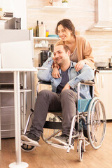 Smiling disabled man in wheelchair and his wife during a video call on laptop in kitchen. Disabled...