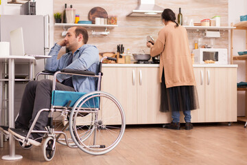 Handicapped man in wheelchair using laptop in kitchen and wife is preparing meal. Disabled paralyzed handicapped man with walking disability integrating after an accident.