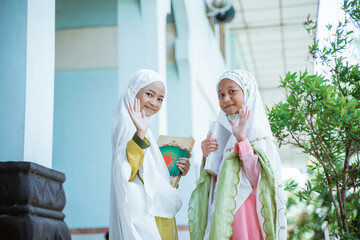 two Asian Muslim girls wearing mukena holding al quran book after salat together and wave hand to camera in the mosque