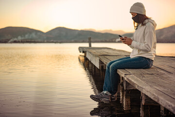 Lonely woman uses her smartphone to follow the latest news on Covid-19 sitting on a pier with...