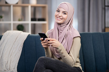 Young attractive smiling muslim woman, holding smartphone in hands, sitting on sofa couch, relaxing in living room at home, and looking at camera during leisure time.