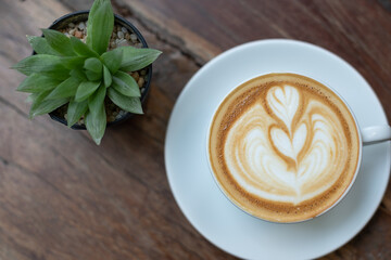 A cup of coffee with heart pattern in a white cup on wooden background with small green tree.
