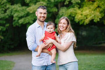 Fototapeta premium Family day. Caucasian smiling mother and father with baby daughter in park. Happy family mom, dad and little girl together outdoor on summer day. Authentic lifestyle real people.