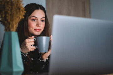 Young caucasian woman with brunette hair resting at home in cozy atmosphere, drinking tea sitting in front laptop and looking on the screen.