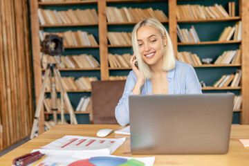 Caucasian female freelancer is having a conversation with colleague, discussing project. Successful business woman is using smartphone, working remotely from home office, sitting at the desk