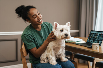 Smiling black woman stroking her dog while sitting at home kitchen