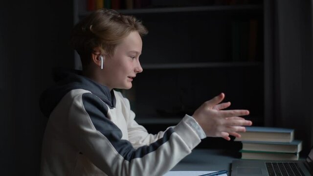 Side view of pupil boy in earphone talking with teacher during online lesson using laptop computer, smiling and gesturing with hands sitting at desk near window at home. Tracking shot in slow motion.