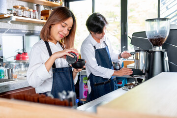 Barista woman or coffee maker pour cream or ingradiant to the cup and stand beside of co-worker man in coffee shop. Concept of happy working with small business and sustainable together.