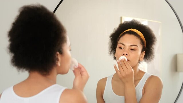 Black african american woman holding cotton pad disk cleaning face skin with cleanser tonic. Skin care remove make up purify lotion in front mirror at home.
