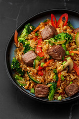 Asian rice with teriyaki beef and vegetables: zucchini, broccoli, pepper, mushrooms, carrot, onion and sesame seeds. Dish isolated in a blue bowl, close-up on a black marble background. Asian cuisine.