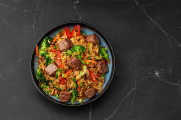 Asian rice with teriyaki beef and vegetables: zucchini, broccoli, pepper, mushrooms, carrot, onion and sesame seeds. Dish isolated in a blue bowl, close-up on a black marble background. Asian cuisine.