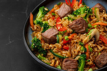 Asian rice with teriyaki beef and vegetables: zucchini, broccoli, pepper, mushrooms, carrot, onion and sesame seeds. Dish isolated in a blue bowl, close-up on a black marble background. Asian cuisine.