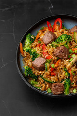 Asian rice with teriyaki beef and vegetables: zucchini, broccoli, pepper, mushrooms, carrot, onion and sesame seeds. Dish isolated in a blue bowl, close-up on a black marble background. Asian cuisine.