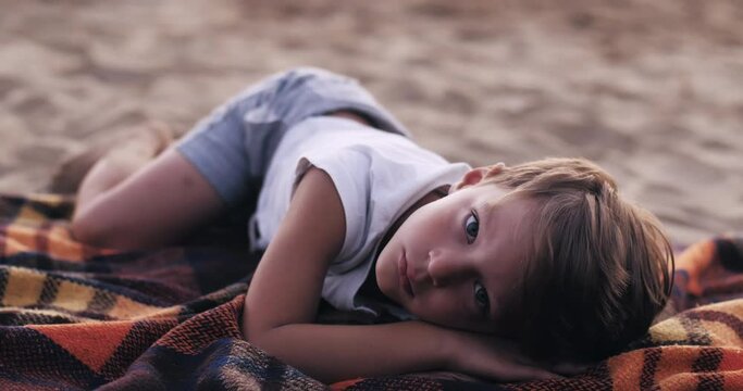 Close-up portrait of cute happy little Caucasian boy child lying down relaxing, posing on sandy sea beach slow motion.