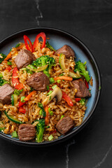 Asian rice with teriyaki beef and vegetables: zucchini, broccoli, pepper, mushrooms, carrot, onion and sesame seeds. Dish isolated in a blue bowl, close-up on a black marble background. Asian cuisine.