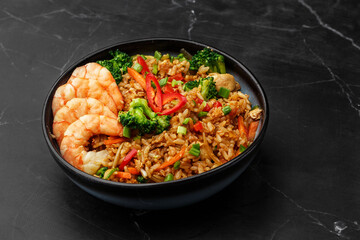 Asian rice with shrimps and vegetables: zucchini, broccoli, pepper, mushrooms, carrot, onion and sesame seeds. Dish isolated in a blue bowl, close-up on a black marble background. Asian cuisine.