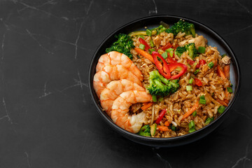Asian rice with shrimps and vegetables: zucchini, broccoli, pepper, mushrooms, carrot, onion and sesame seeds. Dish isolated in a blue bowl, close-up on a black marble background. Asian cuisine.
