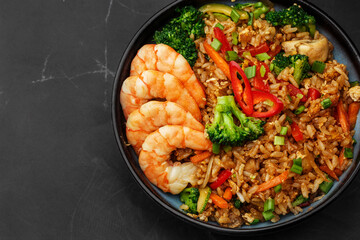 Asian rice with shrimps and vegetables: zucchini, broccoli, pepper, mushrooms, carrot, onion and sesame seeds. Dish isolated in a blue bowl, close-up on a black marble background. Asian cuisine.