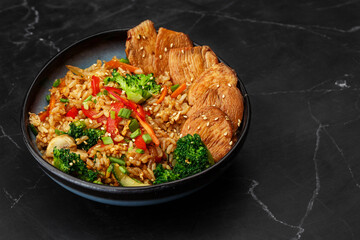 Asian rice with teriyaki chicken and vegetables: zucchini, broccoli, red pepper, mushrooms, carrot, spring onion. Dish isolated in a blue bowl, close-up on a black marble background. Asian cuisine.