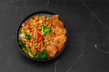 Asian rice with teriyaki chicken and vegetables: zucchini, broccoli, red pepper, mushrooms, carrot, spring onion. Dish isolated in a blue bowl, close-up on a black marble background. Asian cuisine.