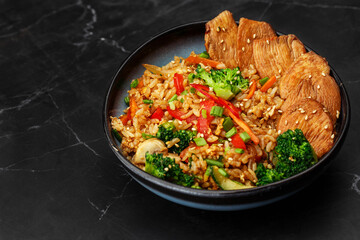 Asian rice with teriyaki chicken and vegetables: zucchini, broccoli, red pepper, mushrooms, carrot, spring onion. Dish isolated in a blue bowl, close-up on a black marble background. Asian cuisine.