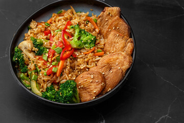 Asian rice with teriyaki chicken and vegetables: zucchini, broccoli, red pepper, mushrooms, carrot, spring onion. Dish isolated in a blue bowl, close-up on a black marble background. Asian cuisine.