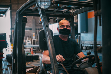 An adult man is seen driving an industrial forklift with a mask