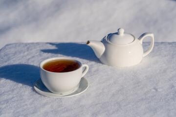 White cup of hot tea and teapot on a bed of snow and white background, close up