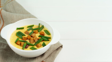 Cheese soup with croutons and green onions in a white plate on a white wooden background.
