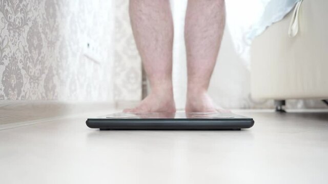 Close-up of a slender man with bare legs taking a step on the scale. checks your weight after a workout at home. Many register weight and fat percentage on digital weight, healthy lifestyle.