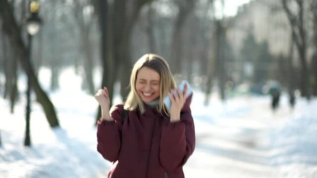 Young woman is very happy to read good news on mobile phone while walking in the city park. Woman jumping and dancing happily