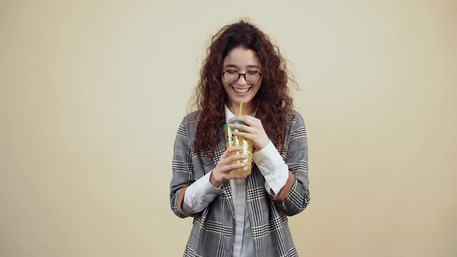 The young woman with curly hair laughs out loud, while she holds in her hand a yellow glass with fresh juice. Young hipster in gray jacket and white shirt, with glasses posing isolated on beige