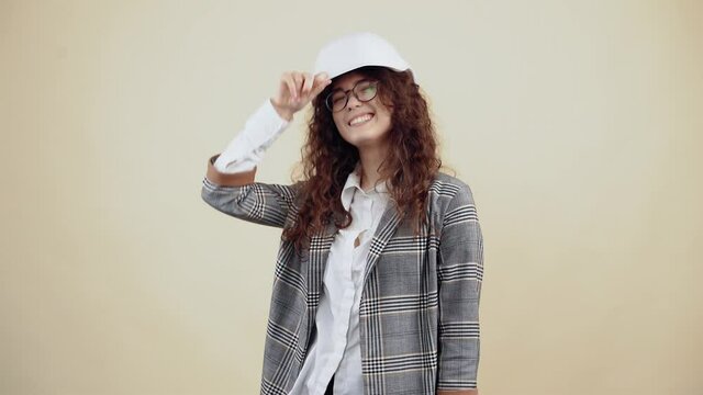 The young woman with curly hair wears a white engineer's helmet, looks around and smiles and shows her tongue. Young hipster in gray jacket and white shirt, with glasses posing isolated on beige
