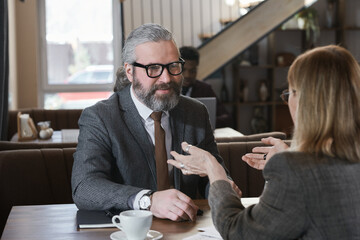 Mature bearded businessman in eyeglasses talking to businesswoman during business lunch in the restaurant