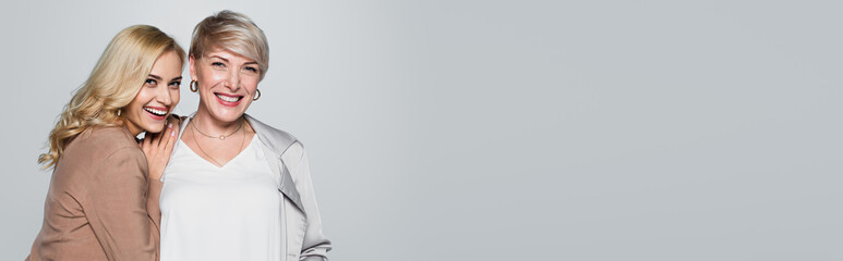 joyful young woman leaning on shoulder of stylish mother isolated on grey, banner
