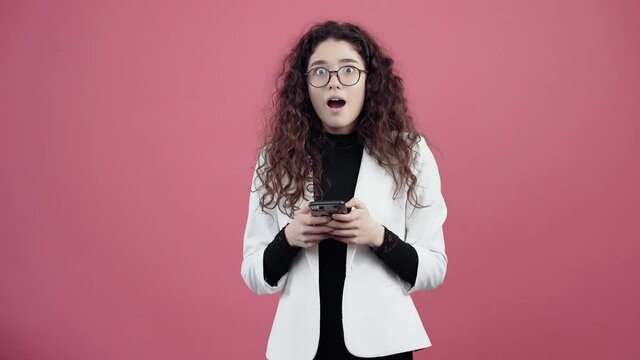 The young woman shocked with curly hair with the phone in her hand looks at the camera with her mouth open, she can't believe it. Young hipster in white jacket and black shirt, with glasses posing