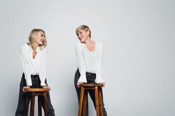Obraz premium joyful mother and daughter smiling at each other while posing near high chairs on grey