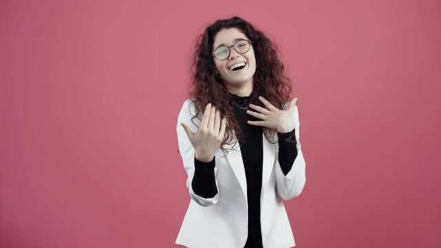 The young adult with curly hair gestures with her hands in front of her, smiling ironically. Young hipster in white jacket and black shirt, with glasses posing isolated on pink background in the