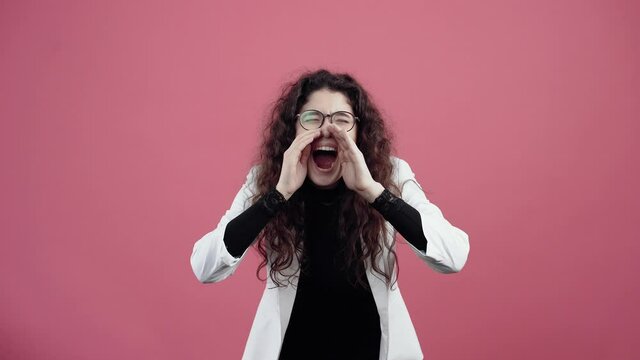 Agitated young woman with curly hair, screams with her hands to her mouth bent forward, with a cascading cure. Young hipster in white jacket and black shirt, with glasses posing isolated on pink