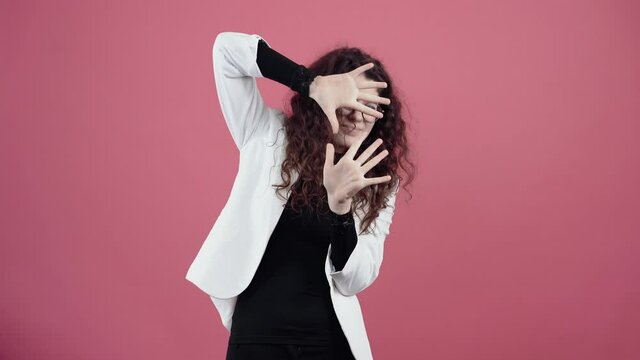 Frightened young woman with curly hair, she keeps her hands in cotton wool hiding behind her palms. Young hipster in white jacket and black shirt, with glasses posing isolated on pink background in