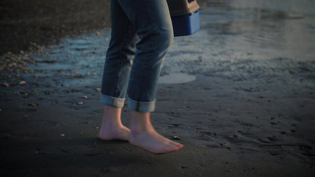 Barefoot man walking on black sand beach with fisherman box in hand, slow motion