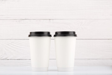 Two white tall disposable coffe cups on white table against white wooden background