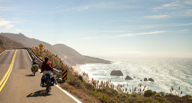 Two cross country bikers pedal down the Pacific Coast Highway with the crashing surf of the Pacific in the background. 