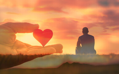 Young man sitting watching the sunset, hand holding heart. Love and nature concept. 