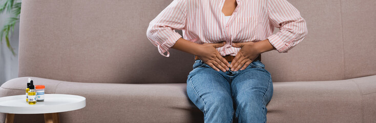 partial view of african american woman suffering from abdominal pain while sitting on sofa in living room, banner