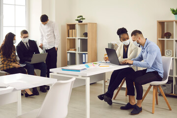 Diverse interracial employee colleague wearing protective face mask to prevent coronavirus COVID-19 spreading working using laptop in office after lockdown. Business lifestyle, cooperation, coworking