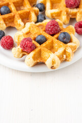 Top view of waffles with raspberries and blueberries, on plate, selective focus, on white table, in vertical, with copy space