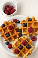 Aerial view of three waffles with raspberries and blueberries, selective focus, on white table with bowl of raspberries, vertical, with copy space