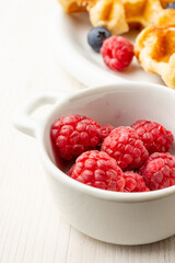 Top view of bowl with raspberries, plate with waffles with raspberries and blueberries on white plate, selective focus, vertical