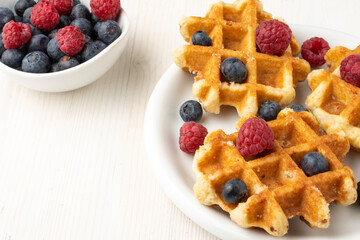 Aerial view of white plate with waffles with raspberries and blueberries, bowl with raspberries and blueberries, selective focus, on white table, horizontal